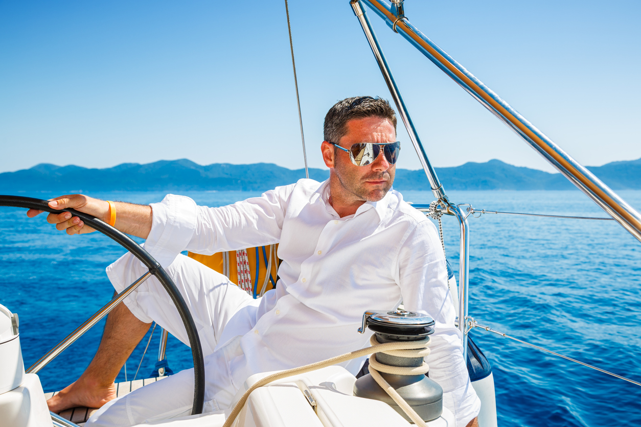  A man in white pants and shirt is steering a sailboat with the ocean and mountains in the background. (Specialized coverage options for small businesses)